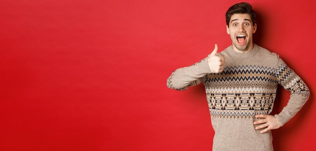 Retrato de hombre atractivo emocionado en suéter de navidad que muestra el pulgar hacia arriba y se ve asombrado recomendando algo bueno publicitando productos relacionados con la navidad