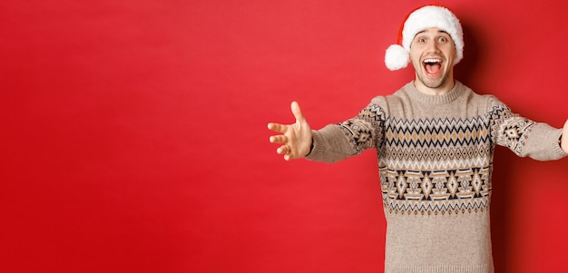 Retrato de hombre atractivo feliz en suéter de navidad y sombrero de santa que alcanza las manos para regalo quiere ...