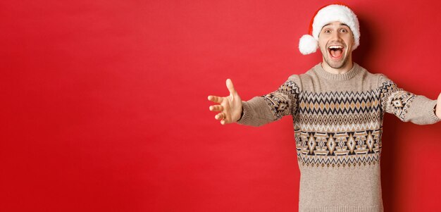 Retrato de hombre atractivo feliz en suéter de navidad y sombrero de santa que alcanza las manos para regalo quiere ...