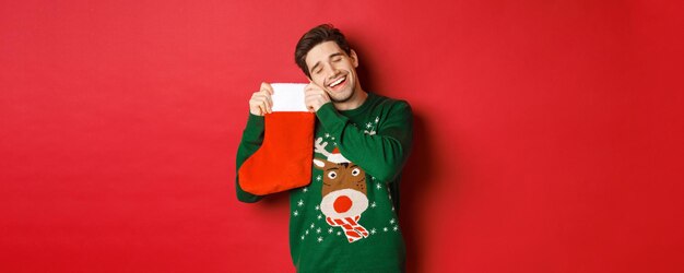 Retrato de hombre atractivo feliz en suéter verde, mirando encantado de recibir regalos en calcetines navideños, de pie sobre fondo rojo.