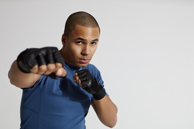 Retrato de hombre atractivo joven de raza mixta con la cabeza rapada haciendo ejercicio en el gimnasio, de pie en la pared blanca, apuntando con el puño bombeado a la cámara, lanzando un puñetazo. Concepto de boxeo, kickboxing y artes marciales.