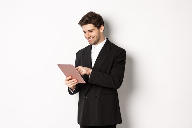 Retrato de hombre atractivo en traje de moda, mirando tableta digital y sonriendo, compras en línea, de pie sobre fondo blanco.