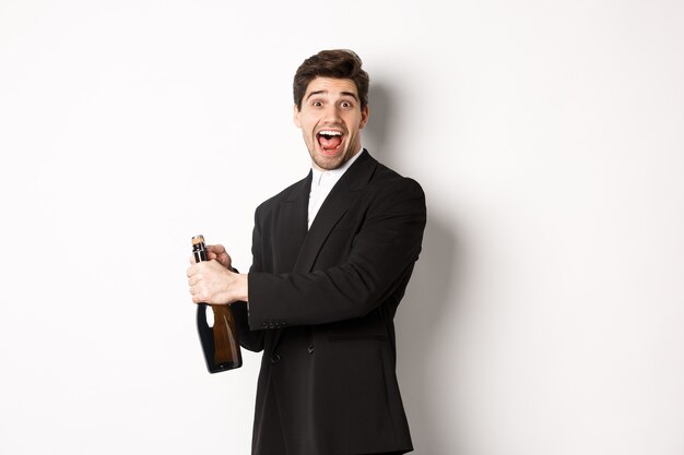 Retrato de hombre atractivo en traje negro, guiñando un ojo a la cámara y abriendo una botella de champán, celebrando el año nuevo, de pie contra el fondo blanco.