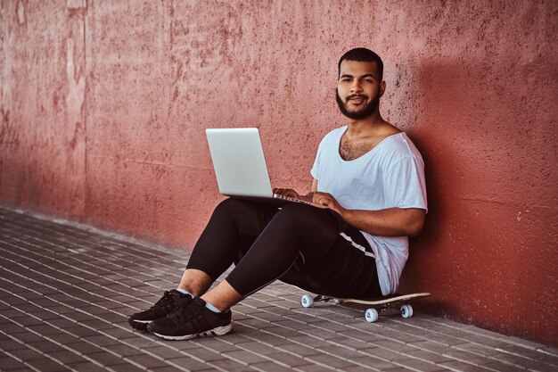 Retrato de un hombre barbudo afroamericano sonriente vestido con una camisa blanca y pantalones cortos deportivos sostiene una laptop mientras se sienta en una patineta debajo de un puente, mirando una cámara.