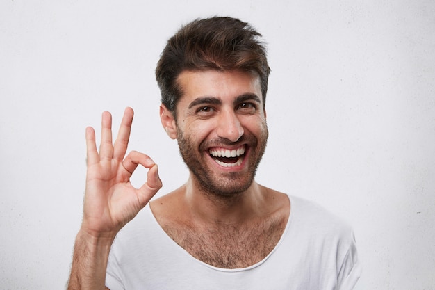 Retrato de hombre barbudo alegre con peinado elegante mostrando signo ok expresando su acuerdo. Joven empresario guapo mostrando su éxito y regocijándose de su triunfo en el trabajo gesticulando