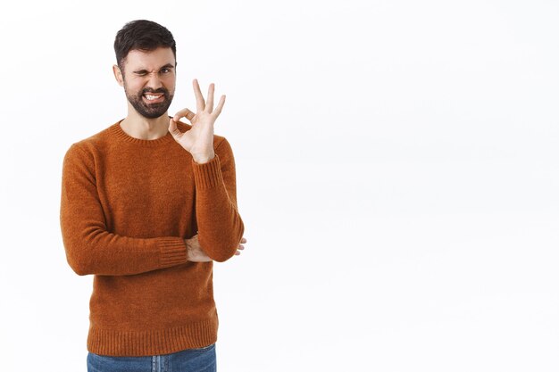 Retrato de hombre barbudo asertivo y guapo, asegura que la calidad es la mejor, muestra un signo de bien, guiña un ojo y sonríe asegurando, teniendo la confianza de recomendar algo, de pie complacido en la pared blanca