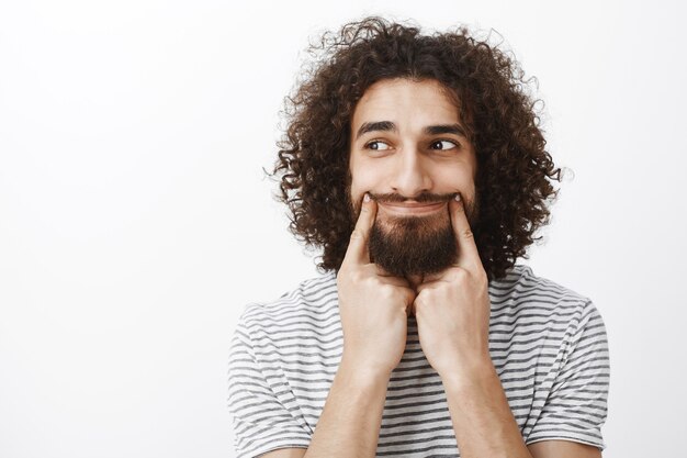 Retrato de hombre barbudo atractivo molesto estresado con cabello rizado, tirando de la sonrisa con el dedo índice y mirando a la izquierda con expresiones tristes, aburrido