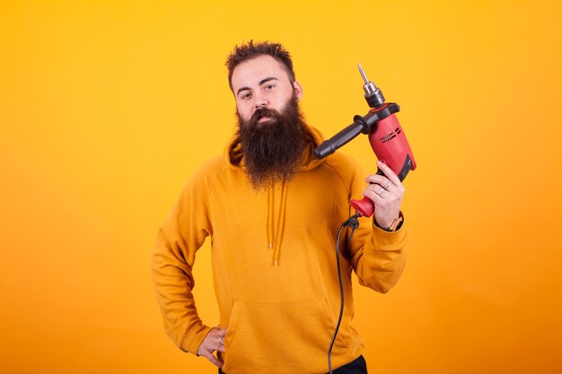 Retrato de hombre barbudo con capucha amarilla sosteniendo un taladro eléctrico rojo y mirando a la cámara sobre fundamento amarillo. Trabajador de sexo masculino. Hombre confiado.