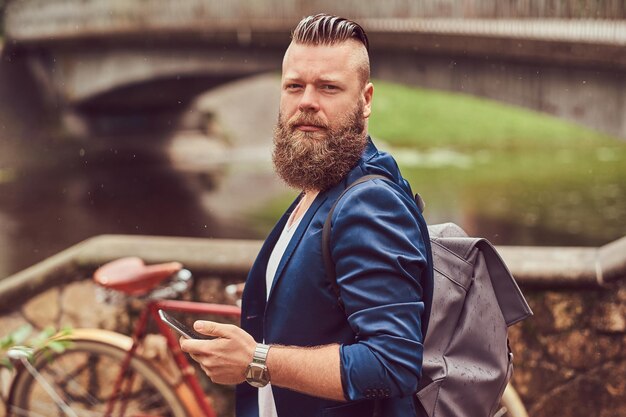 Retrato de un hombre barbudo con un corte de pelo vestido con ropa informal con una mochila, parado en un parque de la ciudad, usando un smartphone.