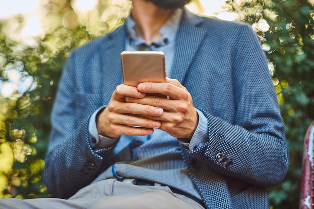 Retrato de un hombre barbudo con un elegante corte de pelo con ropa informal, escribe un mensaje por teléfono mientras se sienta en un banco en un parque de la ciudad.