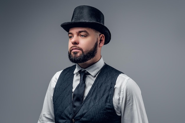 Retrato de hombre barbudo elegante vestido con un chaleco y un sombrero de cilindro aislado sobre fondo gris.