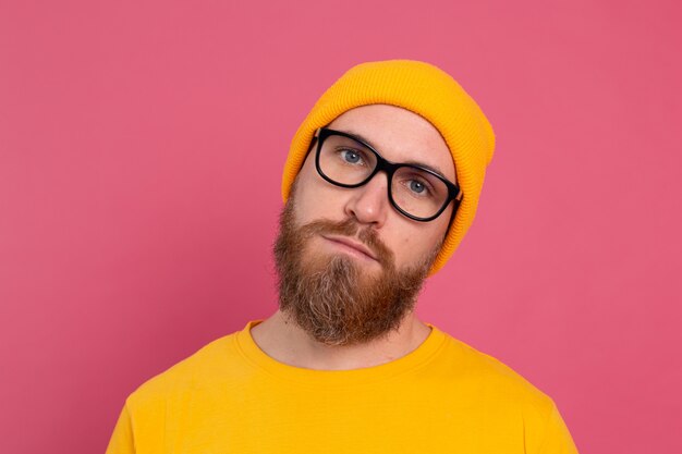Retrato de hombre barbudo europeo guapo elegante con sombrero casual camisa amarilla y gafas sobre fondo rosa