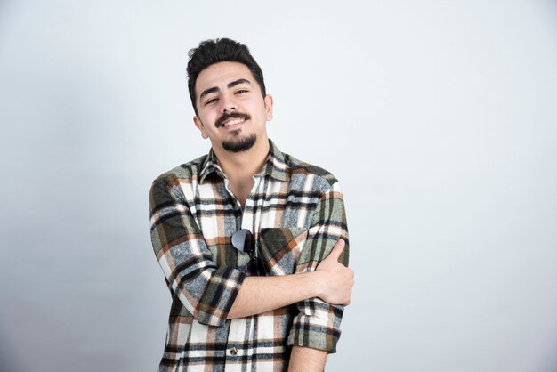 Retrato de hombre barbudo con gafas de pie sobre una pared blanca.