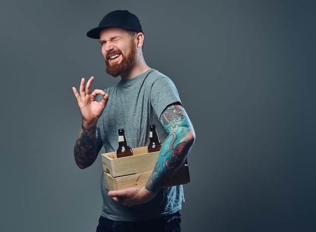 Retrato de un hombre barbudo con una gorra con tatuajes en los brazos sostiene una caja de madera con botellas de cerveza.