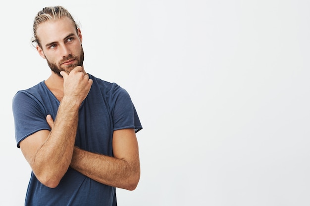 Retrato de hombre barbudo hermoso con elegante peinado y ropa mirando a un lado y pensando