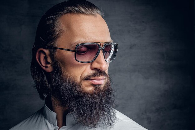 Retrato de un hombre barbudo de lujo con gafas de sol, vestido con camisa blanca sobre fondo gris.