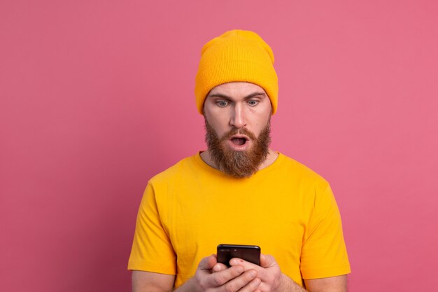 Retrato de hombre barbudo maduro conmocionado preocupado jadeando infeliz sosteniendo smartphone leyendo un mensaje extraño e inquietante en rosa