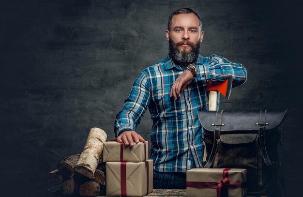 Retrato de hombre barbudo de mediana edad sostiene un hacha y está parado cerca de una mesa con cajas de regalo de Navidad y leña sobre fondo gris.