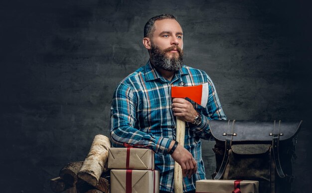 Retrato de hombre barbudo de mediana edad sostiene un hacha y está parado cerca de una mesa con cajas de regalo de Navidad y leña sobre fondo gris.