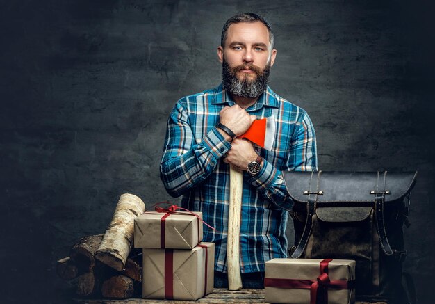 Retrato de hombre barbudo de mediana edad sostiene un hacha y está parado cerca de una mesa con cajas de regalo de navidad y leña sobre fondo gris.