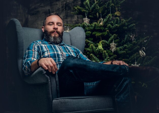 Retrato de hombre barbudo de mediana edad vestido con camisa de franela a cuadros y jeans sentado en una silla sobre fondo decorado de Navidad.