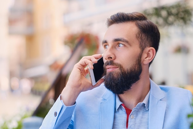 Retrato de hombre barbudo de moda en una chaqueta azul habla por teléfono inteligente.