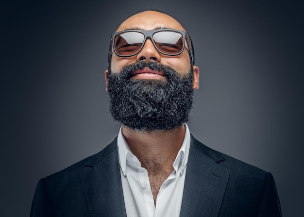 Retrato de un hombre barbudo de moda con traje y gafas de sol aislado en un fondo gris.