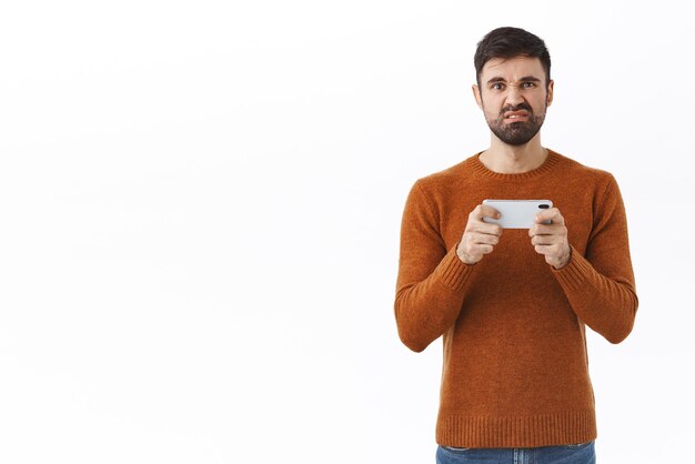 Retrato de hombre barbudo molesto y cabreado que falla la misión en el juego de teléfono inteligente haciendo muecas irritado no puede pasar el nivel difícil jugando en el teléfono móvil de fondo blanco