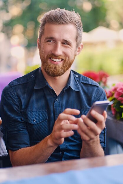 Retrato de hombre barbudo pelirrojo positivo usando un teléfono inteligente.