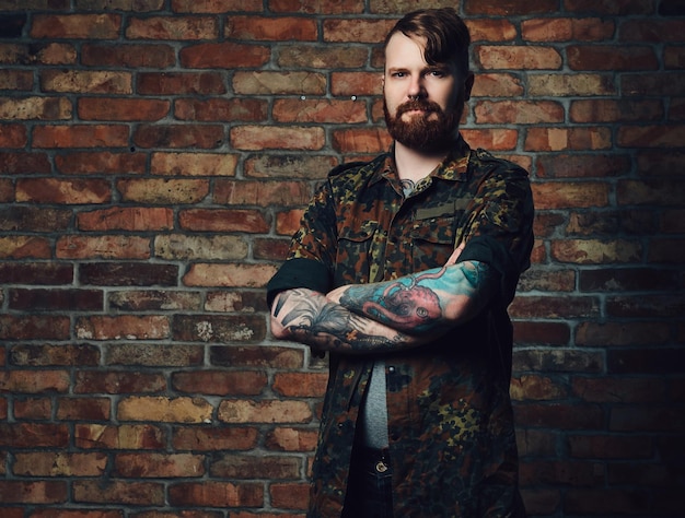 Foto gratuita retrato de hombre barbudo pelirrojo con tatuajes en los brazos, vestido con una chaqueta militar sobre la pared de un ladrillo.