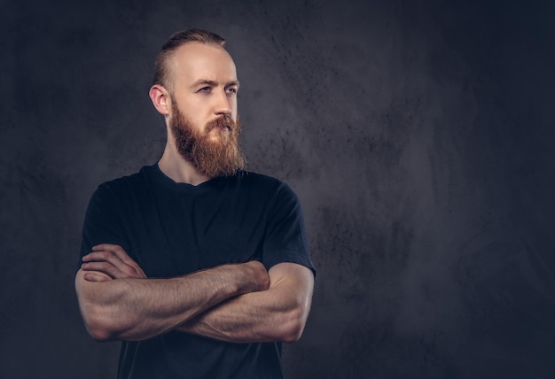 Foto gratuita retrato de un hombre barbudo pelirrojo vestido con una camiseta negra de pie con los brazos cruzados. aislado en el fondo oscuro con textura.