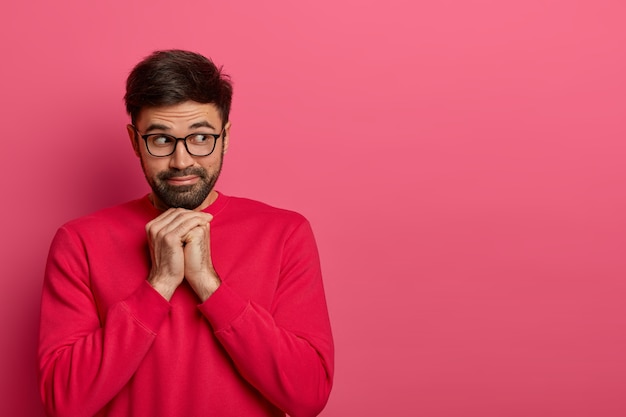 El retrato de un hombre barbudo pensativo mira a un lado, mantiene las manos debajo de la barbilla, aparta la mirada con interés, usa un jersey informal brillante y gafas, piensa en algo. Monocromo. Copie el espacio a un lado para el texto