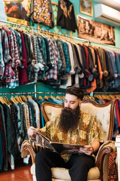 Retrato de un hombre barbudo que se sienta en la silla antigua del brazo que mira la revista en tienda de ropa