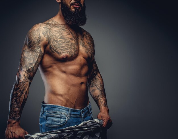 Retrato de hombre barbudo tatuado en jeans y torso desnudo sobre fondo gris.