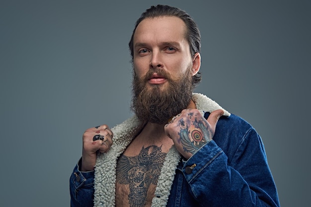 Foto gratuita retrato de hombre barbudo con tatuajes en los brazos y el pecho con una chaqueta vaquera sobre fondo gris.