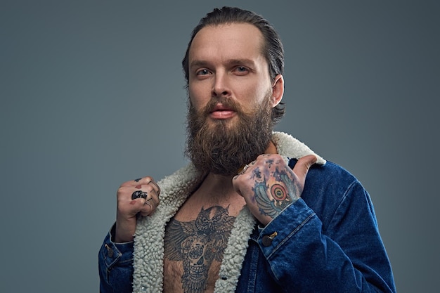 Retrato de hombre barbudo con tatuajes en los brazos y el pecho con una chaqueta vaquera sobre fondo gris.