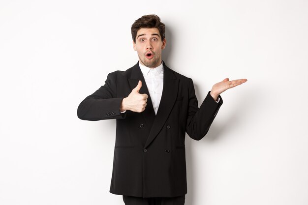 Retrato de un hombre barbudo en traje formal, mostrando el pulgar hacia arriba y sosteniendo el producto en la mano sobre el espacio de la copia en blanco, recomendando el producto, de pie sobre el fondo blanco
