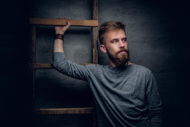 Foto gratuita retrato de un hombre barbudo urbano posando cerca de una vieja escalera vintage sobre fondo gris.