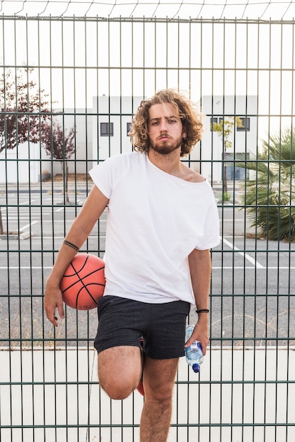 Retrato de un hombre con una botella de baloncesto y agua de pie contra la valla