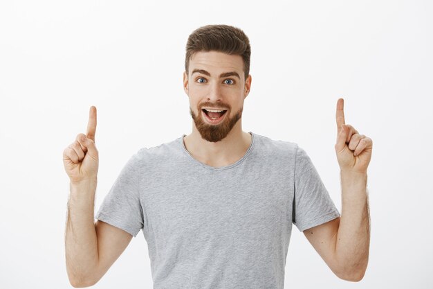 Retrato de hombre brunet saliente entusiasta y emocionante con barba en camiseta gris casual levantando las manos apuntando hacia arriba y discutiendo el increíble espacio de copia hacia arriba posando contra la pared blanca