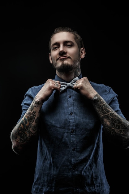 Retrato de un hombre con camisa azul con pajarita gris y tatuajes en las manos. Aislado en negro.
