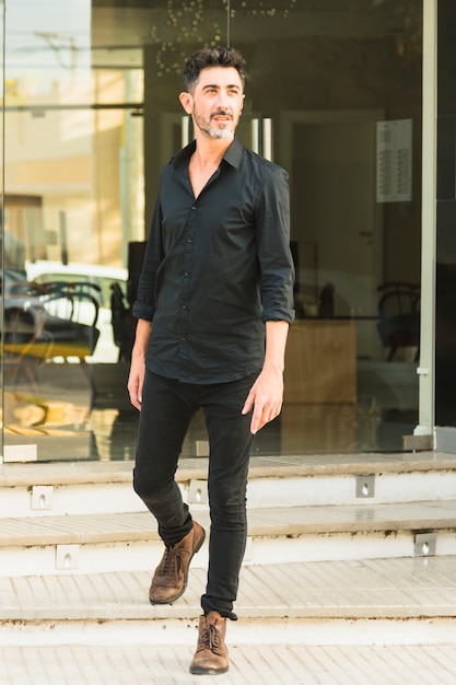 Foto gratuita retrato de un hombre en camisa negra y pantalones vaqueros de pie delante de la puerta de cristal