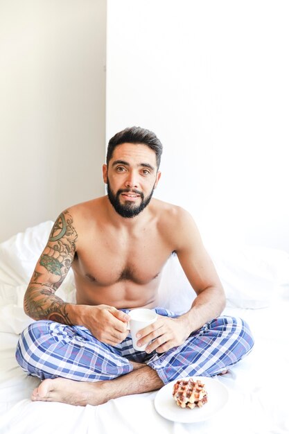 Retrato de un hombre sin camisa sentado en la cama con una taza de café y waffle en un plato