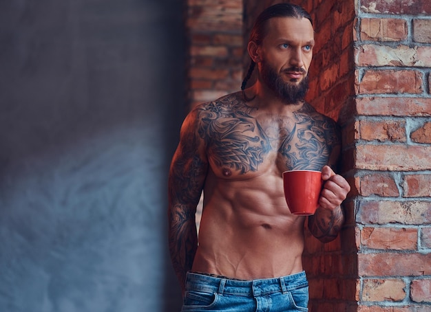 Foto gratuita retrato de un hombre sin camisa tatuado con un elegante corte de pelo y barba, bebe café por la mañana, apoyado contra una pared de ladrillos en una habitación.