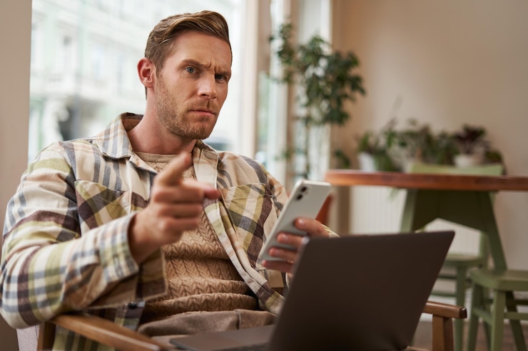 Foto gratuito retrato de un hombre con cara seria sentado en un café con una computadora portátil y un teléfono inteligente apuntando a usted con