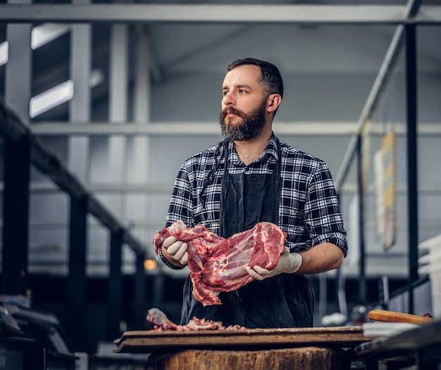 Retrato de un hombre de carne barbudo vestido con una camisa de lana sostiene carne fresca cortada en un mercado.