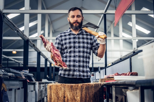 Retrato de un hombre de carne barbudo vestido con una camisa de lana sostiene un hacha y carne fresca cortada.