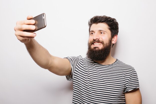 Retrato de un hombre casual alegre haciendo foto selfie en smartphone aislado en una pared blanca