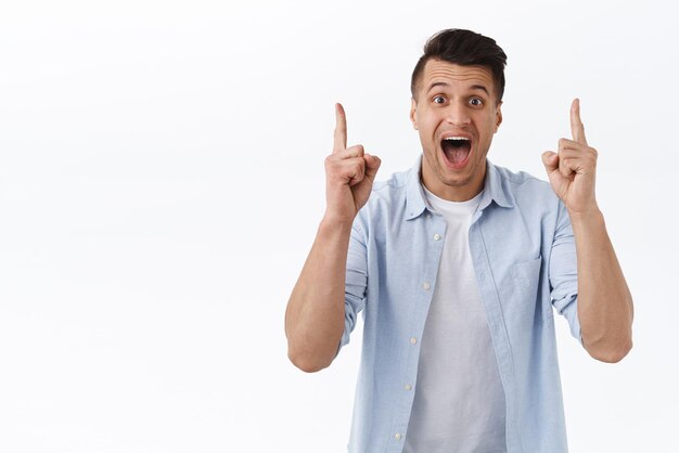 Retrato de un hombre caucásico apuesto, emocionado, alegre e impresionado, señalando con el dedo hacia arriba y sonriendo ampliamente, la cámara muestra una gran oferta, el mejor precio, fondo blanco de pie