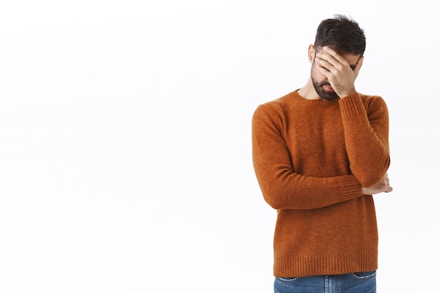 Retrato de hombre caucásico barbudo molesto y cansado, facepalm, siente fatiga y agotamiento después de trabajar hasta altas horas de la noche, suspirando, cerrar los ojos molestos, sentirse incómodo y deprimido, de pie en la pared blanca